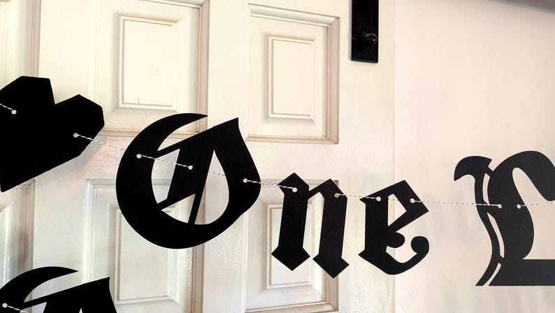 May include: A black paper banner featuring gothic-style lettering that spells out the word One, with a heart-shaped cutout on the left side. The individual letters and the heart are strung together on a thin white string for hanging display.