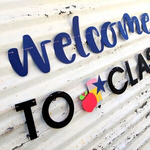 Może przedstawiać: Baner z napisem "Welcome to Class" w niebieskich i czarnych literach. Baner zawiera oł&oacute;wek, gwiazdę i grafikę jabłka. Jest nawleczony na r&oacute;żowy sznurek na białym, zwietrzałym tle.