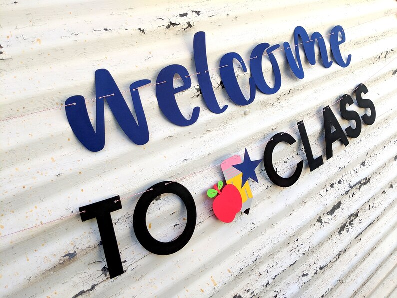 Może przedstawiać: Baner z napisem "Welcome to Class" w niebieskich i czarnych literach. Baner zawiera oł&oacute;wek, gwiazdę i grafikę jabłka. Jest nawleczony na r&oacute;żowy sznurek na białym, zwietrzałym tle.
