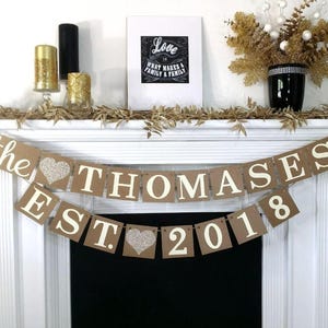 Może przedstawiać: Dekoracyjny baner z napisem "the THOMASES EST 2018" w białych literach na brązowych kwadratach z kartonu. Baner zawiera dwa brokatowe srebrne serca. W tle oprawiony wydruk i świece.