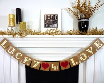 May include: A decorative mantelpiece with a banner that reads "LUCKY IN LOVE." The banner has brown square cards with white letters and red heart accents. The mantel also features candles, a framed print with text, and a black vase with gold foliage.