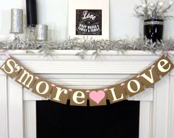 May include: A decorative mantelpiece with a banner that reads "S'more Love" in white letters on brown cardstock. A pink heart is between the words. The mantel also features candles, a framed print, and decorative silver foliage.