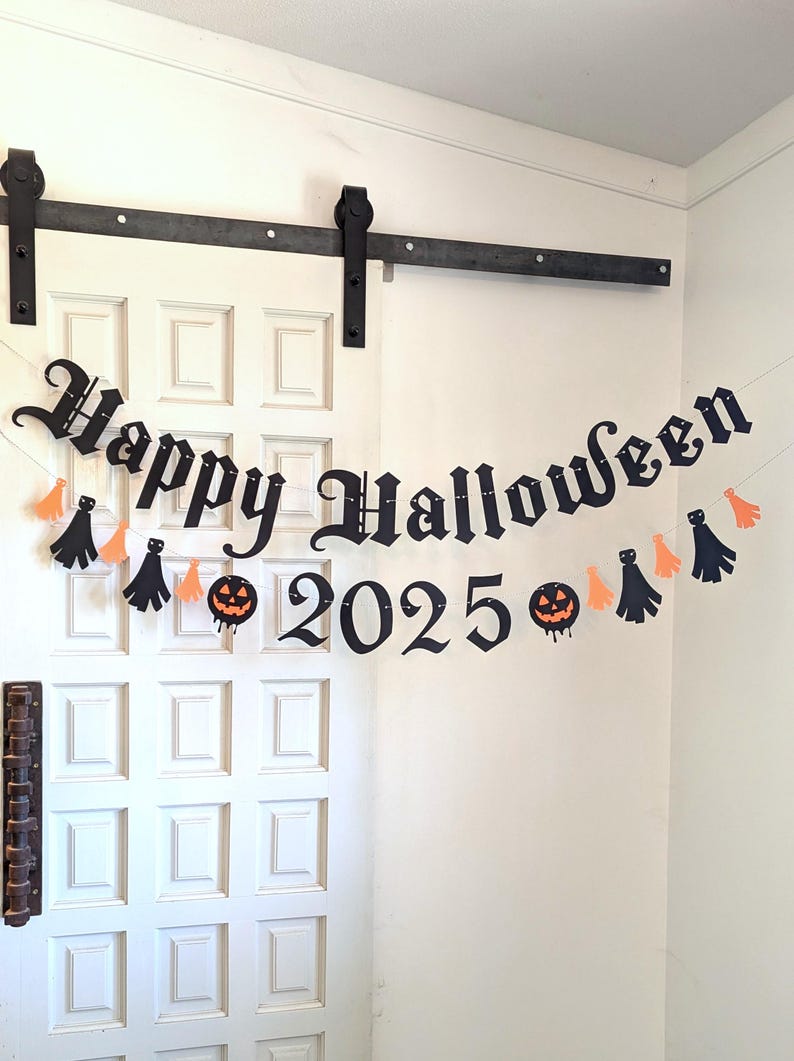 May include: A black Halloween banner with the words "Happy Halloween 2025" in a gothic font, strung across a white door. The banner includes orange and black decorative shapes, including pumpkins and ghosts.