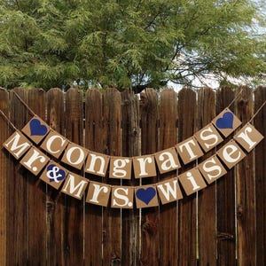 May include: A banner with the words "Congrats Mr. & Mrs. Wiser" in white letters on brown cardboard squares. Blue heart accents are placed between the words. The banner hangs on a wooden fence.