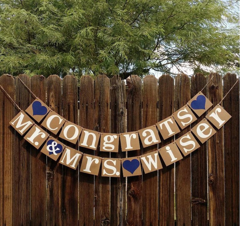 May include: A banner with the words "Congrats Mr. & Mrs. Wiser" in white letters on brown cardboard squares. Blue heart accents are placed between the words. The banner hangs on a wooden fence.