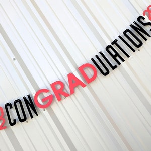 May include: A graduation banner with the words "CONGRATULATIONS 2022" in black and red lettering. The banner is strung diagonally against a white background with vertical lines.