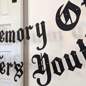 May include: Black banner with the words "In Memory Jennifer's Youth" in a gothic font. The banner is strung on a thin silver wire and hangs in front of a white door and wall.
