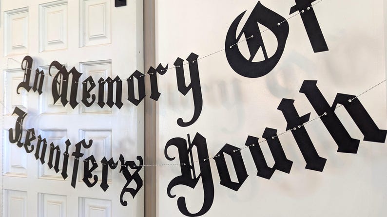 May include: Black banner with the words "In Memory Jennifer's Youth" in a gothic font. The banner is strung on a thin silver wire and hangs in front of a white door and wall.