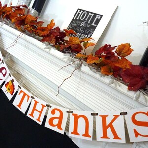 May include: Thanksgiving banner with the words "GIVE THANKS" in orange and burgundy letters. The banner is strung across a white mantel decorated with autumn leaves and a framed sign that reads "HOME is where the heart is".