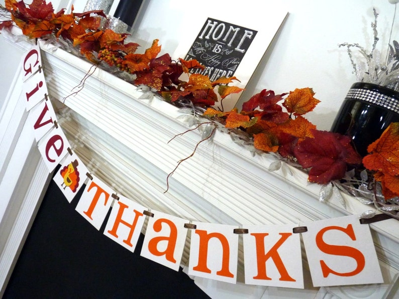May include: Thanksgiving banner with the words "GIVE THANKS" in orange and burgundy letters. The banner is strung across a white mantel decorated with autumn leaves and a framed sign that reads "HOME is where the heart is".