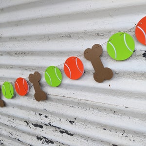 May include: A festive dog-themed banner featuring orange tennis balls, green tennis balls, and brown bone cutouts strung together. The banner is designed with a playful aesthetic, perfect for a pet-themed party or celebration.