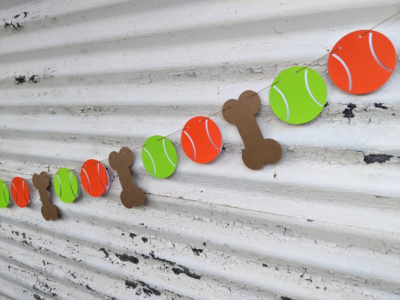 May include: A festive dog-themed banner featuring orange tennis balls, green tennis balls, and brown bone cutouts strung together. The banner is designed with a playful aesthetic, perfect for a pet-themed party or celebration.