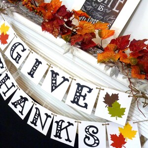 May include: A white banner with the words "GIVE THANKS" in black, decorative lettering. The banner is strung with twine and decorated with colorful fall leaves. Autumnal foliage and a framed sign complete the festive display.