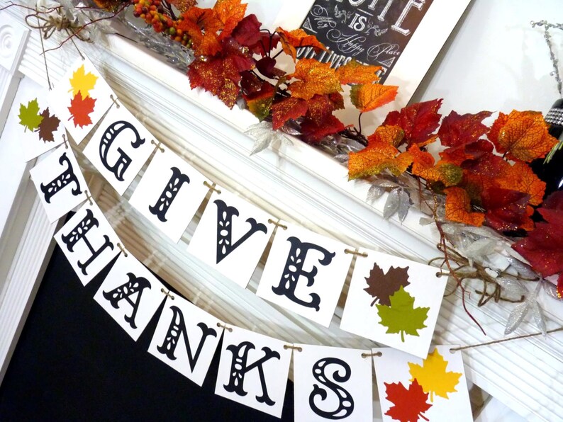 May include: A white banner with the words "GIVE THANKS" in black, decorative lettering. The banner is strung with twine and decorated with colorful fall leaves. Autumnal foliage and a framed sign complete the festive display.