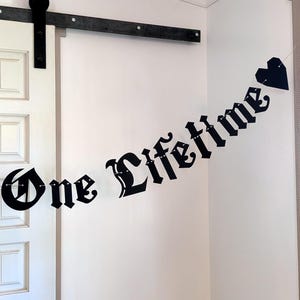 May include: A black banner featuring the text One Lifetime in a gothic font, with a black heart shape at the end. The letters are connected by a thin string, creating a hanging decoration that displays the phrase clearly against a plain wall.