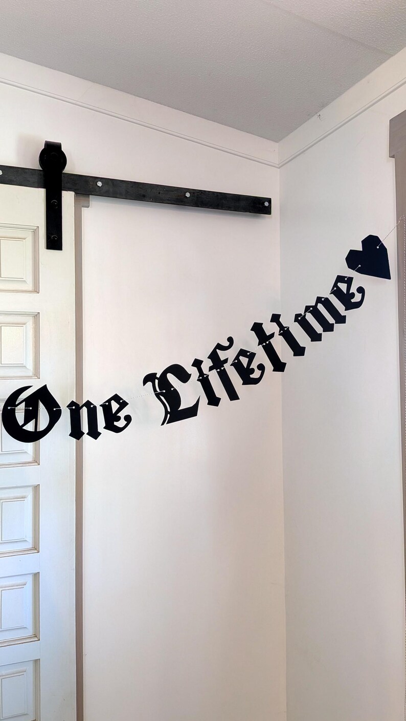 May include: A black banner featuring the text One Lifetime in a gothic font, with a black heart shape at the end. The letters are connected by a thin string, creating a hanging decoration that displays the phrase clearly against a plain wall.