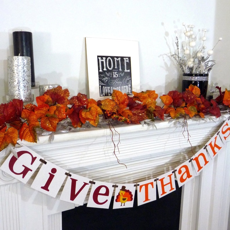 May include: Thanksgiving mantelpiece decoration with a banner reading "Give Thanks" and a turkey illustration. The mantel is decorated with autumn leaves, candles, and a framed sign saying "Home is where love lives here."