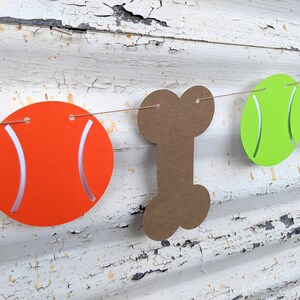 May include: A festive banner featuring orange tennis balls, a brown bone, and a green tennis ball strung together. The orange balls have white curved accents, and the banner is suspended against a distressed white wooden background.
