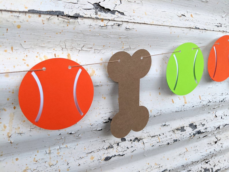 May include: A festive banner featuring orange tennis balls, a brown bone, and a green tennis ball strung together. The orange balls have white curved accents, and the banner is suspended against a distressed white wooden background.