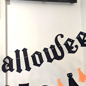 May include: A black Halloween banner with the word "Halloween" in a gothic font. The banner is strung with small lights and includes orange and black decorative cutouts. The banner is hanging in front of a white door.