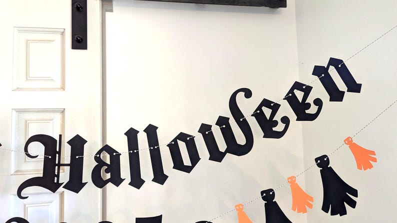 May include: A black Halloween banner with the word "Halloween" in a gothic font. The banner is strung with small lights and includes orange and black decorative cutouts. The banner is hanging in front of a white door.