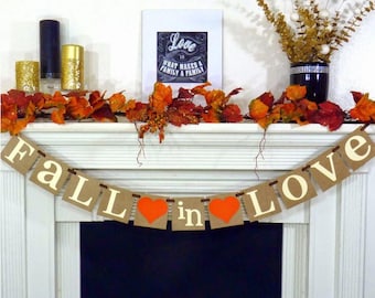 May include: A mantel decorated for fall with a banner that reads "FALL in LOVE" with orange hearts. The mantel also features fall leaves, candles, and a framed print that says "Love is what makes a family a family."