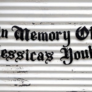 May include: Black metal sign with the words "In Memory Of Jessica's Youth" in a gothic font. The sign is attached to a white, weathered, corrugated metal surface.
