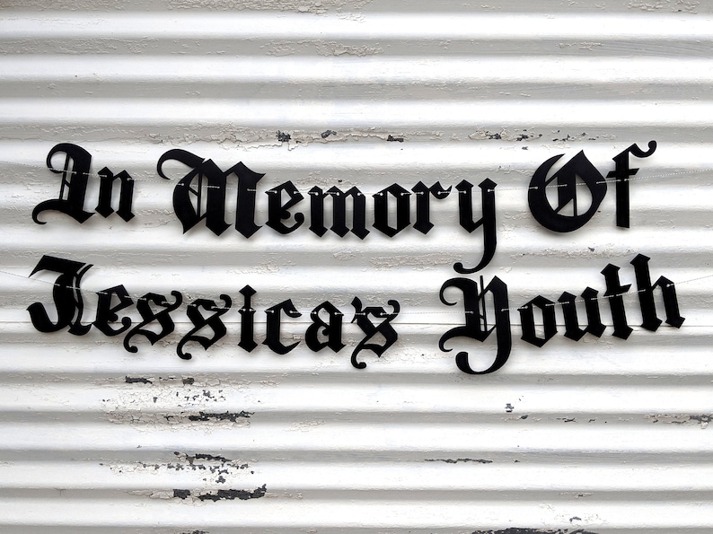 May include: Black metal sign with the words "In Memory Of Jessica's Youth" in a gothic font. The sign is attached to a white, weathered, corrugated metal surface.