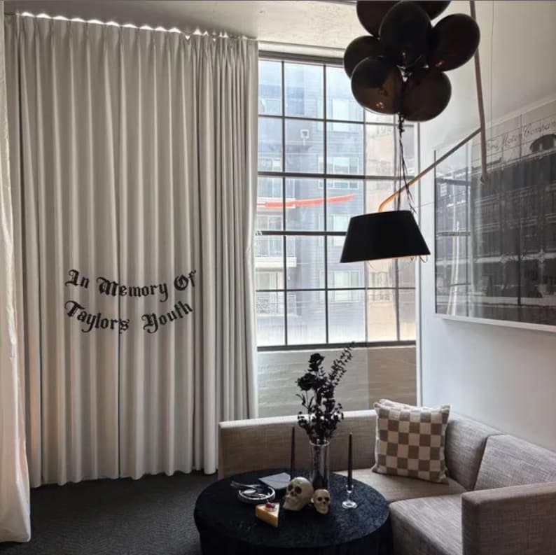 May include: A living room scene with a black round table, a gray sofa, and a large window. The curtains have the text "In Memory Of Taylors Youth". Black balloons and a black lamp hang from the ceiling. Decorative skulls and candles are on the table.
