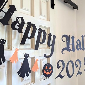 May include: A Halloween banner with the words "Happy Halloween 2025" in black lettering. The banner includes orange and black paper cutouts of ghosts and tassels, strung on a thin white string, and is hanging on a white door.
