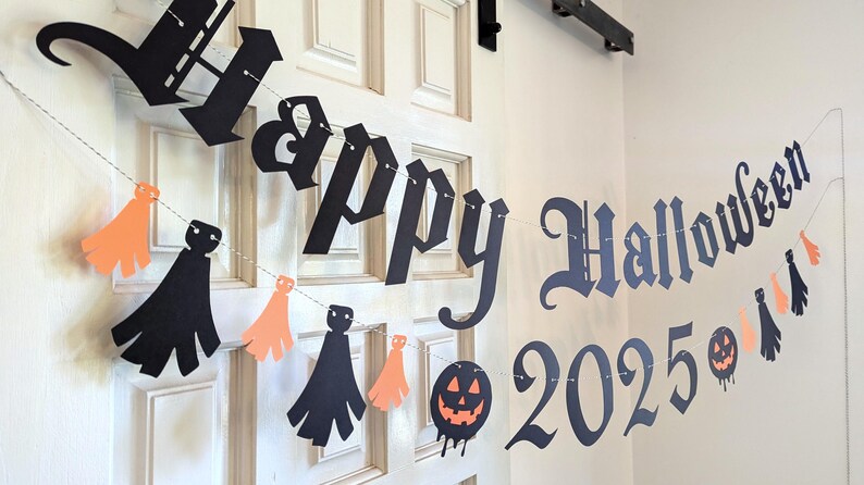 May include: A Halloween banner with the words "Happy Halloween 2025" in black lettering. The banner includes orange and black paper cutouts of ghosts and tassels, strung on a thin white string, and is hanging on a white door.