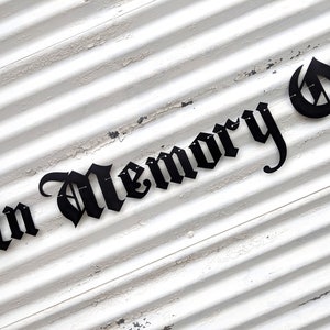 May include: Black metal sign with the words "In Memory Of" in a gothic font. The sign is mounted on a white, weathered, corrugated metal surface.