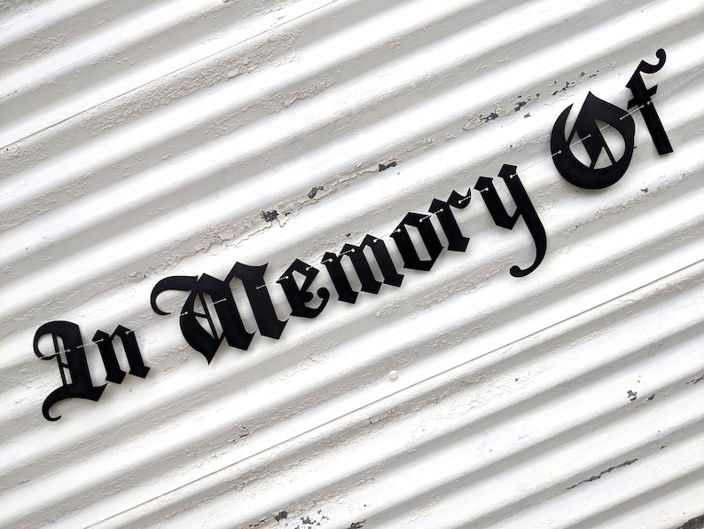 May include: Black metal sign with the words "In Memory Of" in a gothic font. The sign is mounted on a white, weathered, corrugated metal surface.