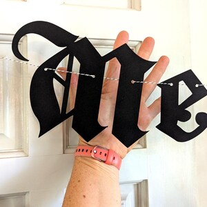 May include: Black banner spelling "In Memoriam" in a gothic font. The banner is strung on a white string and held up against a white door. The banner is made of black paper.