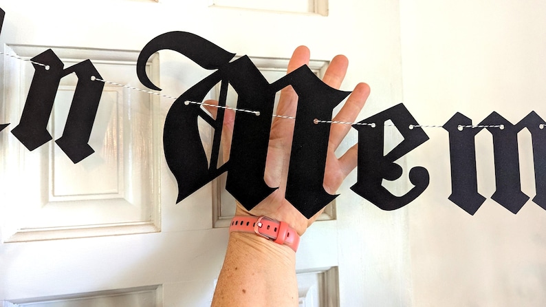 May include: Black banner spelling "In Memoriam" in a gothic font. The banner is strung on a white string and held up against a white door. The banner is made of black paper.