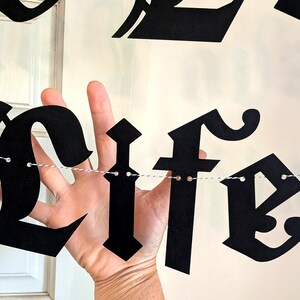May include: A black paper banner featuring decorative gothic-style lettering spelling out the word life. The individual letters are cut from matte black cardstock and are strung together on a thin, white and black twisted twine string.