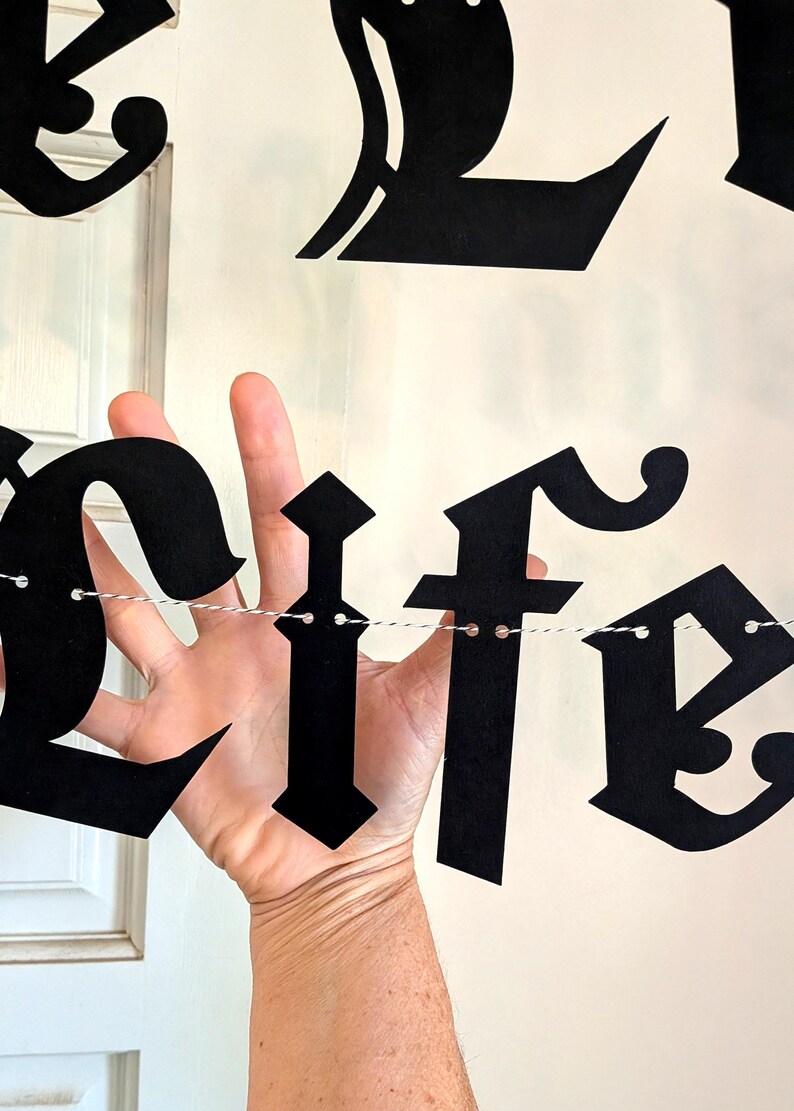May include: A black paper banner featuring decorative gothic-style lettering spelling out the word life. The individual letters are cut from matte black cardstock and are strung together on a thin, white and black twisted twine string.