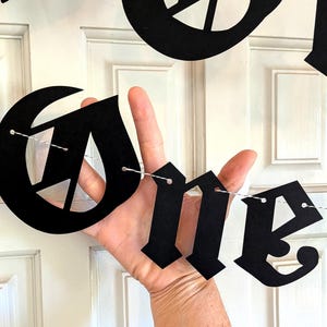 May include: A black banner featuring large, die-cut letters in a gothic-style font spelling out the word ONE. The individual letters are suspended on a thin, white and black twisted string, creating a decorative hanging sign for display.