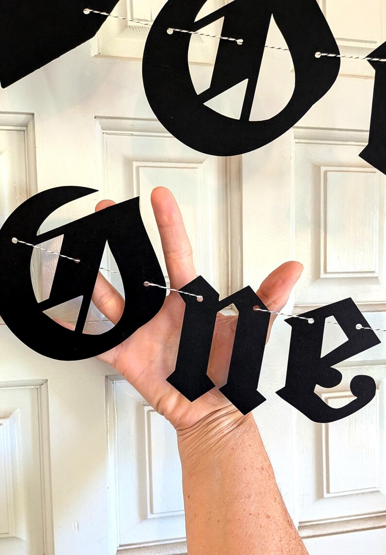 May include: A black banner featuring large, die-cut letters in a gothic-style font spelling out the word ONE. The individual letters are suspended on a thin, white and black twisted string, creating a decorative hanging sign for display.