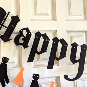 May include: A black paper banner with the word "Happy" in a gothic font is strung across a white door. Below, a string of orange and black paper cutouts of ghosts and capes adds a festive touch, perfect for Halloween.