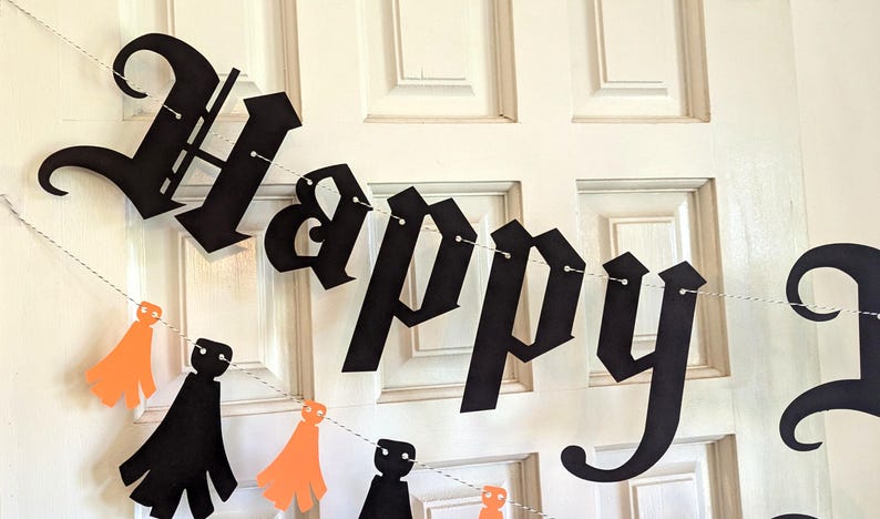 May include: A black paper banner with the word "Happy" in a gothic font is strung across a white door. Below, a string of orange and black paper cutouts of ghosts and capes adds a festive touch, perfect for Halloween.