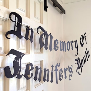 May include: A black paper banner with the words "In Memory of Jennifer's Youth" in a gothic font is strung across a white door. The banner is suspended by a thin white string, creating a decorative memorial.