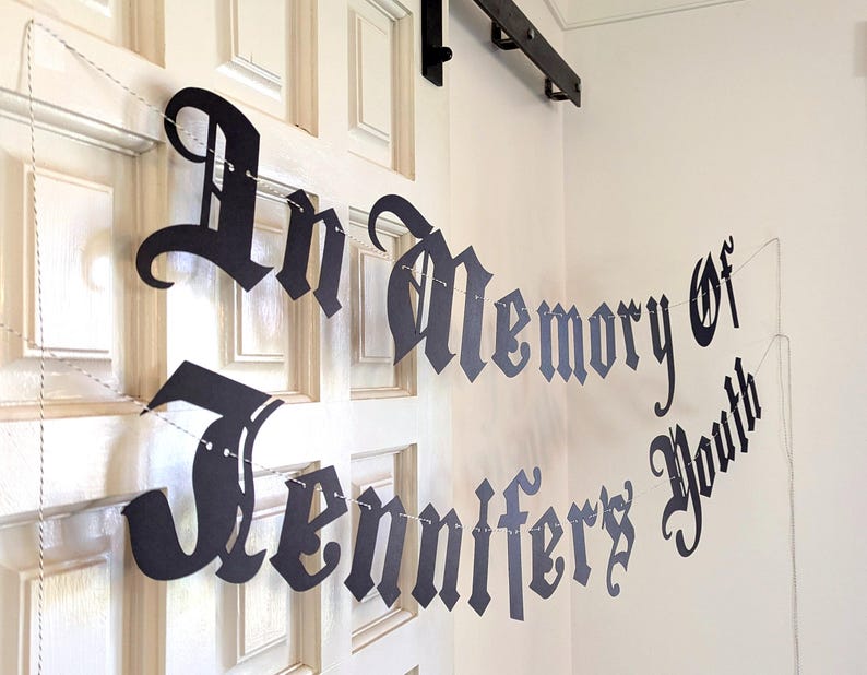 May include: A black paper banner with the words "In Memory of Jennifer's Youth" in a gothic font is strung across a white door. The banner is suspended by a thin white string, creating a decorative memorial.