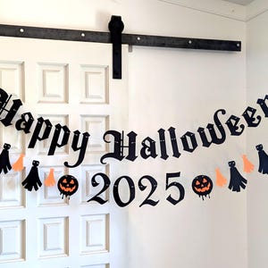 May include: A black banner with the words "Happy Halloween 2025" in a gothic font, strung across a white door. The banner is decorated with orange and black tassels and jack-o'-lanterns, creating a festive Halloween decoration.