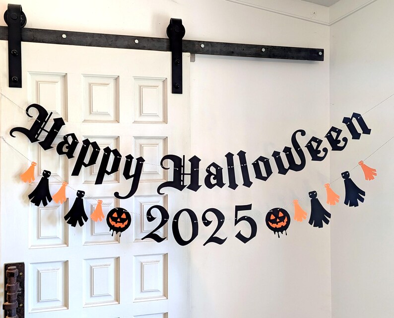 May include: A black banner with the words "Happy Halloween 2025" in a gothic font, strung across a white door. The banner is decorated with orange and black tassels and jack-o'-lanterns, creating a festive Halloween decoration.
