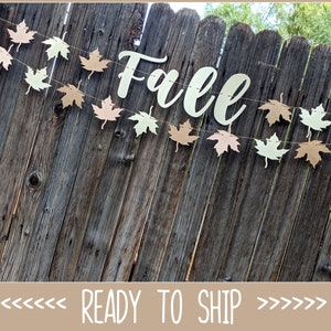 May include: A fall-themed banner with maple leaf cutouts in shades of beige and green, strung across a rustic wooden fence. The word "Fall" is displayed in elegant cursive. The banner is a decorative item for autumn celebrations.