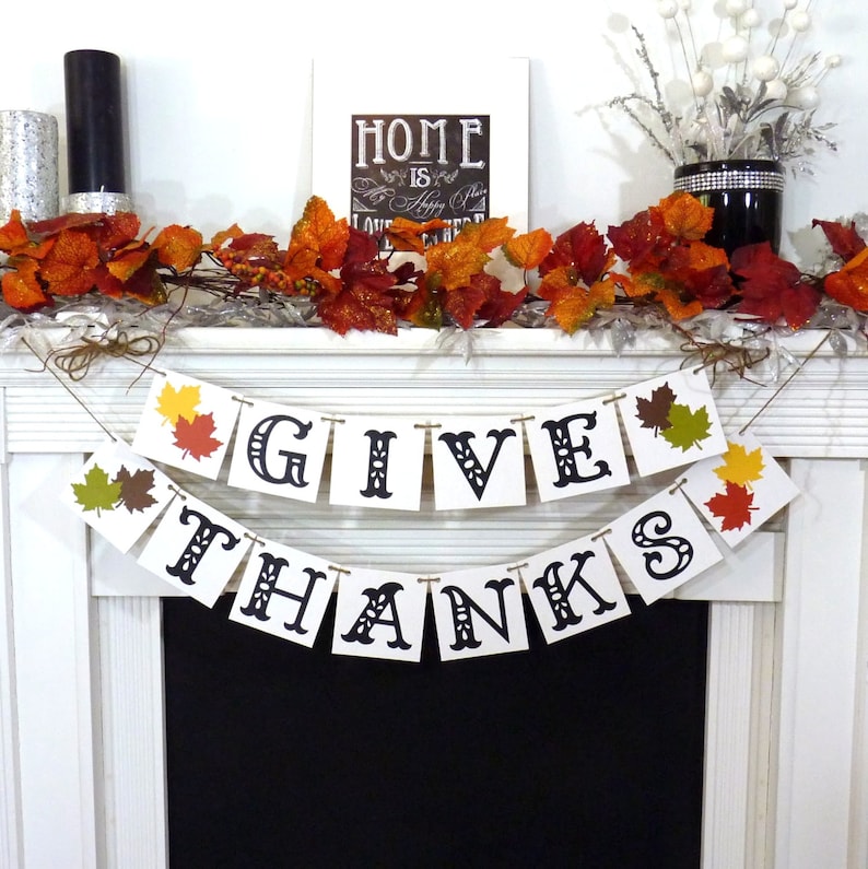 May include: A festive Thanksgiving banner with the words "GIVE THANKS" in black letters. The banner is strung with twine and decorated with colorful fall leaves. A garland of autumn leaves and berries adorns the mantel, with a framed sign that reads "HOME is where happiness starts."