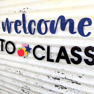 Może przedstawiać: Baner z napisem "welcome to class" w granatowych i czarnych literach. Baner zawiera czerwone jabłko, niebieską gwiazdę i ż&oacute;łty oł&oacute;wek. Baner jest zawieszony na r&oacute;żowym sznurku na białym, zwietrzałym tle.