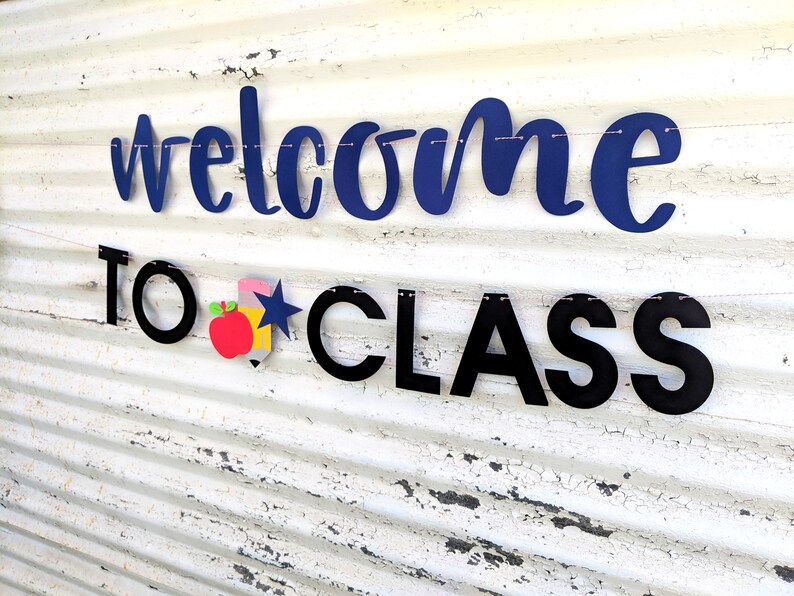 Może przedstawiać: Baner z napisem "welcome to class" w granatowych i czarnych literach. Baner zawiera czerwone jabłko, niebieską gwiazdę i ż&oacute;łty oł&oacute;wek. Baner jest zawieszony na r&oacute;żowym sznurku na białym, zwietrzałym tle.
