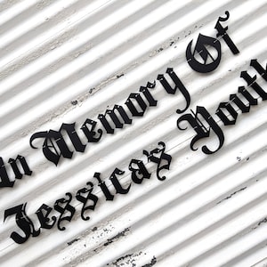 May include: Black metal sign with the words "In Memory Of Jessicaz Youth" in a gothic font. The sign is mounted on a white, weathered, slatted wooden background.